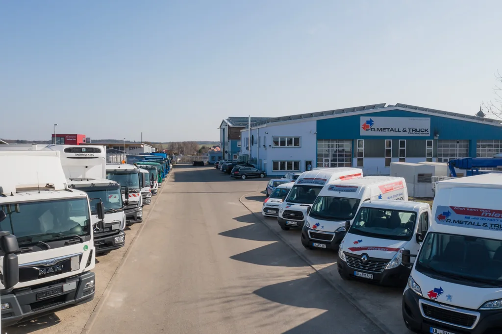 Blick über den Betriebshof mit LKW-Reihe, Transportern und der Halle von R. Metall & Truck Trading.